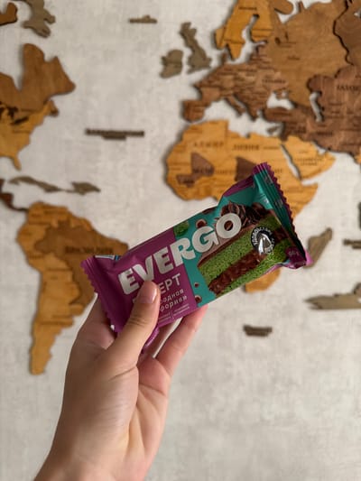 A hand, with light skin and a pale pink manicure, holds a teal and purple wrapper for a chocolate bar against a wooden map of the world. The wrapper features the brand "EVERGO" and an image of a slice of green cake with chocolate filling. Text on the wrapper is in Cyrillic, and some is blurred. The background is a wall with a wooden world map. The provided crops do not offer enough detail to determine the specific location within Semiluki, Russia.
