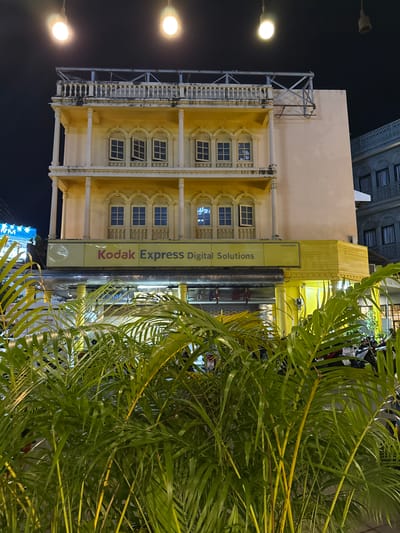 This is a medium shot of a building at night, likely in Phuket City Municipality, Thailand. The building is a pale yellow structure with ornate white detailing around the windows and along the facade. Several windows are visible, some of which are open. A tall, yellow sign with the text "Kodak Express Digital Solutions" is attached to the building. The sign is vertical and appears to be illuminated. In the foreground, lush green palm fronds partially obscure the view of the building. The scene is lit by artificial light sources, including streetlights that cast a bright glow. The sky is dark, indicating it is nighttime. The overall impression is of a commercial establishment in an urban environment.