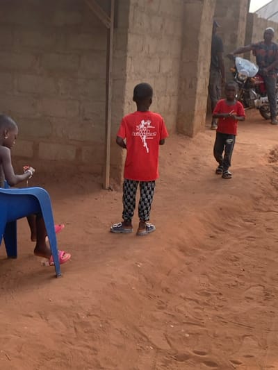 Several children and an adult are outdoors on a dirt ground near a building. One child wears a red shirt with a white design and patterned pants, standing facing away. Another child, also in a red shirt, is standing further away. One child is sitting on a blue plastic chair wearing pink slippers. An adult sits on a motorcycle in the background wearing a patterned shirt.