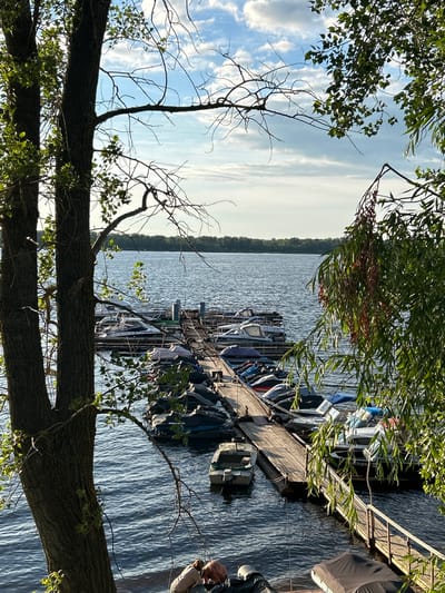 The image depicts a serene lake scene with a wooden dock lined with various boats. Lush green foliage from overhanging trees frames the top and left sides of the shot, casting dappled shadows. The water appears calm with gentle ripples reflecting the bright daylight. In the background, a distant tree line marks the edge of the lake. Several boats, ranging in size and style, are moored to the dock, some covered with tarps. A person can be partially seen on the right side of the dock, tending to the boats. The sky is a mix of blue and white clouds, suggesting a pleasant day. The overall atmosphere is peaceful and idyllic, characteristic of a summer day at a lakeside. There is no visible text in the image.