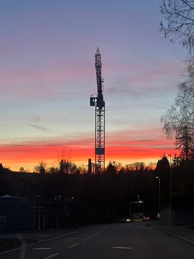 The image shows a construction crane silhouetted against a vibrant sunset in Wetzikon (ZH), Switzerland. The sky displays a gradient of colors from violet to orange, with a band of light-colored clouds above the horizon. A road runs along the right side of the image, with street lights, trees, and buildings visible in the background. The scene captures a construction project during the twilight.