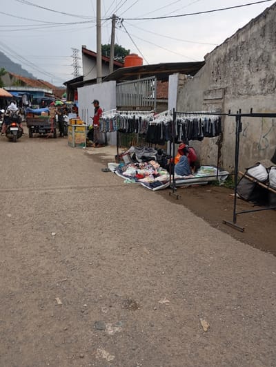 The scene is a street in Kutawaringin, Indonesia, located at latitude -6.974 and longitude 107.52. There are two people sitting on the ground, a man standing next to a rack of clothing, a motorcycle, a small cart, and a variety of other objects. The man is wearing a red shirt and jeans, and the two people sitting on the ground are wearing casual clothing. The ground is covered in loose gravel, and there is a small pile of clothing on the ground in front of the people sitting down. The clothing on the rack appears to be mostly jeans, and there are several other items, such as towels, bags, and shoes, piled on the ground next to the rack. There is a building in the background, and a large metal gate to the left of the clothing rack. The scene is likely a market or a small store.