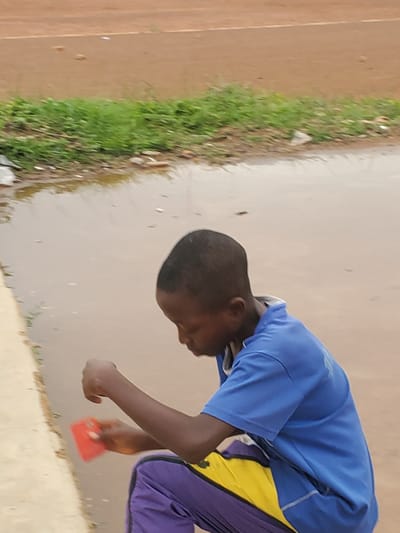 A young boy, likely of Nigerian descent and from Pankshin, Nigeria, is the sole subject in this photograph. He is depicted from the chest down, wearing a bright blue t-shirt with yellow and purple accents on his pants. The boy is bent over, his head down, and is holding a small, rectangular, red object in his right hand, which appears to be a toy or a similar small item. His left hand is also visible, bent at the elbow.
The setting appears to be outdoors on a paved surface, possibly a pathway or road, with a strip of green grass bordering the paved area. The ground surface is a light, neutral color, suggesting it could be concrete or a similar material. Scattered on the grass and the paved area are small specks, which could be debris or dust.
The lighting suggests it is daytime, with no distinct shadows indicating a particular time. The weather seems to be clear, with no visible rain or clouds. The overall mood of the image is neutral, focusing on the boy's activity. There is no visible text in the photograph. The image is oriented sideways.