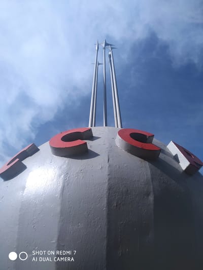 The image features a large, grey, spherical structure with the red letters "TCC" affixed to its surface. Three white, cylindrical poles topped with star-shaped ornaments extend upwards from the top of the sphere. The background is a bright blue sky with scattered white clouds. The words "SHOT ON REDMI 7 AI DUAL CAMERA" appear at the bottom of the frame. This monument is located in the city of Foki, Russia.