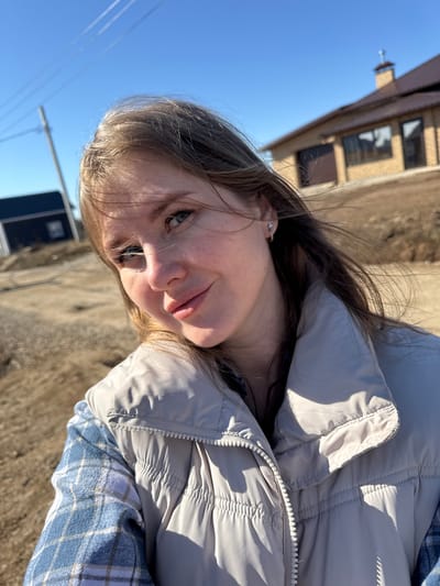 A young woman is posing outdoors in Gornyi (Dvurechenskoe S/P), Russia. She is wearing a light grey puffy jacket over a blue and white plaid shirt. The location appears to be near a building with brown brickwork under a clear blue sky with utility wires. The ground is light brown. The angle suggests the picture was taken from a low vantage point, looking slightly upwards.