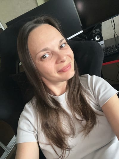 This is a selfie of a young woman with long, brown hair, wearing a light-colored t-shirt. She is looking at the camera with a slight smile and her eyes are open. The lighting suggests it is daytime, and the background is somewhat blurred, indicating an indoor setting. Visible in the background are computer monitors, a keyboard, and a speaker, suggesting she is in a home office or gaming setup. The overall mood of the image appears relaxed and casual. There is no visible text in the image.