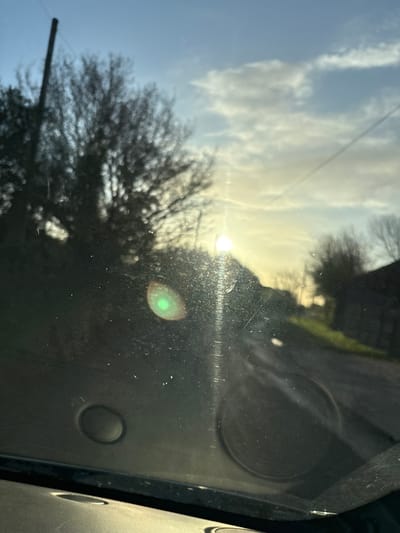 The image captures an exterior scene in Ruyton-XI-Towns, United Kingdom, viewed through the dirty and scratched windshield of a vehicle. The low sun, positioned in the upper right quadrant, causes significant lens flare, visible as a prominent green circular artifact in the lower-center and a vertical streak of light. The sky above is clear blue with scattered white clouds, transitioning to a bright golden hue near the horizon due to the sunlight.
In the immediate foreground, within the vehicle, the top edge of a textured dashboard is visible. A circular, black recessed object is present on the lower-left, while a circular speaker grille is visible through the windshield on the lower-right. The windshield itself exhibits numerous dust particles, smudges, and fine scratches.
Outside the vehicle, the environment is out of focus due to the focal point being on the windshield. On the far left, a dark, vertical pole is silhouetted against the sky, partially obscuring a large, bare-branched deciduous tree. A narrow, unpaved or gravel road or track extends into the background on the right, flanked by areas of green grass. Further into the background, indistinct, dark building structures and additional bare trees are silhouetted against the bright sky. No individuals, animals, or active subjects are visible in the scene.