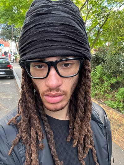 A man with brown dreadlocks, wearing a black head wrap and black-rimmed glasses, is the primary subject of the image. He is positioned outdoors, with greenery and trees visible in the background. A street is also visible, including a black car with a license plate partially visible, reading "FQ-0P". The man appears to be standing near a brick structure. The overall location is likely in an urban area, specifically Les Ulis, France.