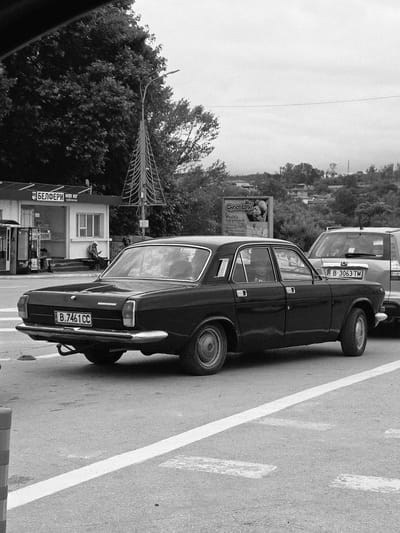 In Beloslav, Bulgaria, a black sedan is parked on an asphalt road, facing away from the viewer. The car's license plate reads "B.7461 CC". To the right of the sedan, a silver SUV is also parked, its license plate visible as "B.3063 TM". Behind the vehicles, a building with signs in Cyrillic is visible, appearing to be a kiosk or small shop. A street light with a unique, geometric design stands near the building. In the background, rolling hills covered in trees and houses suggest a semi-urban or suburban environment under an overcast sky. Two individuals are seated on a bench outside the building. The scene appears to be a street scene during daylight.