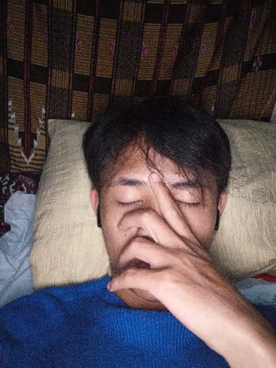 A person reclines against a pillow, eyes closed, and hand covering the mouth and nose. The individual has short, dark hair and wears wireless earbuds. A blue sweater is visible. The background features a dark brown and tan textured fabric, possibly a curtain or blanket. Lighting is dim. The composition is a close-up, emphasizing the subject's face and hand.