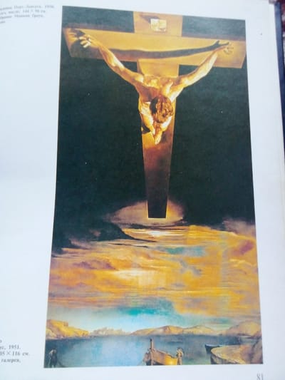 The image is a painting depicting a religious scene, specifically the crucifixion of Jesus Christ. The figure of Christ is rendered in a golden hue, with his arms outstretched and body contorted. He is suspended on a large wooden cross against a dark, almost black, background. Below the cross, the painting transitions to a surreal landscape. A body of water, colored in vibrant blues and turquoise, stretches across the foreground, with a rocky, mountainous coastline visible in the distance. The sky above the water is filled with dramatic, swirling clouds in shades of orange, yellow, and pink, illuminated by an unseen light source. In the water, there are several small boats, some with figures standing in them or on the shore. The painting appears to be titled "Corpus Hypercubus" or "Christ of Saint John of the Cross" by Salvador Dalí, painted in 1951. The text on the left side of the image, partially obscured, indicates that the painting is in the collection of the Metropolitan Museum of Art. There is no indication that this artwork is located in Zhmerynka, Ukraine; instead, it is associated with a museum in New York City.