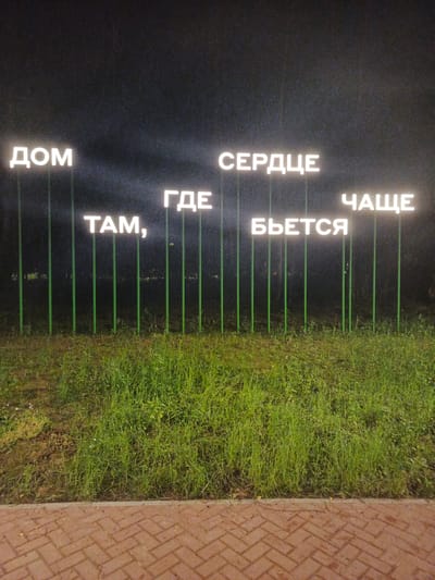 The image depicts a nighttime scene in Radkevshchina, Russia, featuring illuminated text displayed on vertical poles. The text is in Russian and appears to be a quote or phrase. The phrase reads, "ДОМ ТАМ, ГДЕ СЕРДЦЕ БЬЕТСЯ ЧАЩЕ" which translates to "Home is where the heart beats more often."
The illuminated letters are white and stand out against the dark background, suggesting it is nighttime. The poles supporting the letters are green and arranged in a line across a grassy area. In the foreground, there is a brick pathway. The grass appears lush and green, indicating a possibly temperate climate, and there is some moisture on it, suggesting recent rain or dew.
There are no people visible in the image. The focus is solely on the text installation and its surroundings. The lighting creates a somewhat reflective and atmospheric mood. The overall impression is one of a public art installation or a decorative feature, possibly in a park or outdoor public space. The clear night sky and the artificial lighting of the text are the dominant elements of the scene.