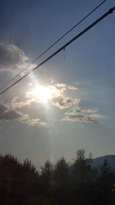 The image depicts a bright sky with scattered clouds and the sun shining through, casting light rays. Two power lines run diagonally across the upper portion of the frame. Below the sky, a silhouetted line of trees forms the lower part of the scene, with a hint of a hill or mountain in the far distance. The overall impression is of a daytime outdoor scene, possibly taken in Liptovský Michal, Slovakia, given the context provided. There are no people or animals visible in the image. The scene is static and there are no discernible actions or interactions taking place. The environment appears to be natural, with vegetation and a hilly landscape.