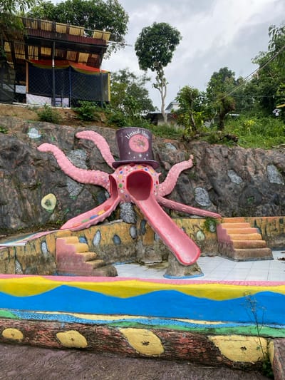 The image shows a children's playground in the form of a pink octopus wearing a black top hat with a "Umbul Neso" inscription. The octopus is a slide with a pink, curved ramp. The playground is situated on a rocky hillside, adorned with colorful painted designs. The hillside is surrounded by lush greenery and trees. The image was taken in Kecamatan Manyaran, Indonesia, located at latitude -7.826 and longitude 110.816. A small concrete structure with a metal fence is visible in the upper left corner of the image. A partially visible house with a white facade can be seen in the background to the right of the slide. The ground is a mixture of concrete and dirt. The sky is overcast with grey clouds.