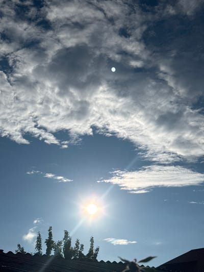 The image captures a bright, sunny day with a clear blue sky dotted with various types of clouds. The sun, a prominent and radiant orb, is visible in the upper right quadrant, casting a strong glare and sunbeams. To the left of the sun, a small, pale circular object is also present in the sky, potentially the moon or a lens flare artifact.
The sky is filled with a mix of fluffy white cumulus clouds and wispy, elongated cirrus clouds. Some of the cumulus clouds appear dense and dark, suggesting they may be a bit heavier or further away, while others are bright and illuminated by the sun.
In the lower right corner, the edge of a dark, corrugated roof is visible, and above it, the dark silhouette of tree branches with green foliage is seen. These elements suggest an outdoor setting, possibly in a rural or suburban area, with a building nearby.
The overall scene depicts a pleasant, clear day with good visibility. The strong sunlight indicates it is likely daytime, possibly late morning or early afternoon. The location context of Soloneshnoye, Russia, places the scene in a geographical region. No text is visible in the image.