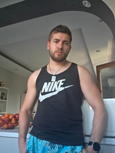 An adult male subject is depicted standing indoors, likely within a domestic setting in Yagodovo, Bulgaria. The subject has short, dark brown hair and a beard, light skin tone, and blue eyes, maintaining a neutral facial expression. He is wearing a black tank top with a white "NIKE" logo printed on the chest, light blue patterned shorts, a silver-colored chain necklace with a rectangular pendant, and a dark-colored smartwatch on his left wrist.
To the subject's left, a white countertop holds a black wire fruit bowl containing a variety of red and orange fruits, including pomegranates, apples, and oranges. Behind the countertop, a white shelving unit is visible, displaying several objects and books. To the subject's right, a white appliance is partially visible, alongside a wooden-framed opening, possibly a doorway or window, leading to another area. The ceiling is white with dark grey/black curved and straight architectural elements, incorporating recessed circular light fixtures. The walls are light-colored. The subject's hands are mostly obscured by his shorts and body.
