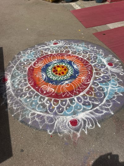 A colorful rangoli, a traditional Indian folk art, is depicted on an asphalt surface in Puttaparthi, India. The intricate design features concentric circles of various colors, including red, orange, blue, and green, accented with white chalk patterns. The rangoli appears to be placed on a ground surface with some red mats visible in the background, suggesting an outdoor setting. The sunlight casts shadows on the ground, indicating it is daytime. There are no people or animals in the frame, and the focus is entirely on the artistic creation.