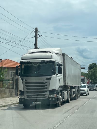 A medium shot of a large, white Scania R440 truck traveling on a road. The license plate reads "CB 5037 AB". The truck has a box trailer attached. Overhead electrical wires and a utility pole are visible. A low building with an orange tile roof and a fenced yard is to the left of the truck. Additional vehicles are stopped ahead of the truck in the distance. The image appears to be taken in Tsar Kaloyan, Bulgaria.