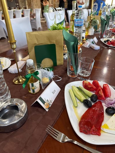 This is a photograph of a table setting at an outdoor event, possibly a celebration or reception, in Sredets, Bulgaria. The table is covered with a brown tablecloth. On the table are various items, including a gold candle holder with a gold candle, a small glass bottle with a green ribbon and a label that reads "70", a white gift bag with a gold chocolate, a white folded card with a green border and a floral design, green napkins, and several glasses. A white plate with a salad containing tomatoes, cucumbers, olives, red onion, and a piece of red pepper and white cheese is also visible. In the background, there are white-covered chairs and people dressed in formal attire. The overall impression is one of a festive gathering.