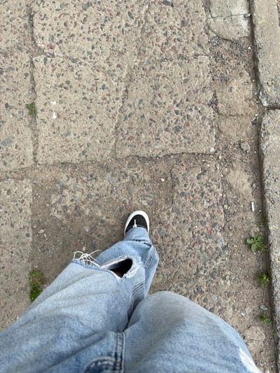A person's legs and feet, clad in ripped light-wash denim jeans and black and white sneakers, are visible from a high-angle perspective. The individual is standing on a cracked and weathered concrete sidewalk, characterized by its gritty texture and embedded stones. The environment appears to be an outdoor urban setting, potentially in the city of Mahilyow, Belarus, given the typical construction materials and condition of public walkways found in the region. Small patches of green vegetation are growing through cracks in the concrete. The lighting suggests it is daytime.