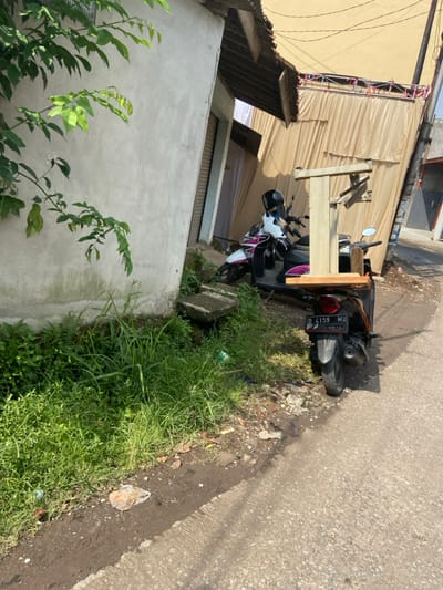 The image depicts a narrow street lined with concrete and paved with asphalt. A light-colored concrete building is visible on the left side of the street. There is a white and pink motorcycle with a license plate that reads "D 4159 MU" parked on the right side of the street. The motorcycle is next to a beige table with a wooden top and a metal frame, positioned perpendicular to the street. There is a second motorcycle, a white scooter with a black seat, partially obscured by the beige table. Lush, green foliage is visible on the left side of the street. The ground is covered with loose gravel and dirt. The image was taken in Margaasih, Indonesia, at coordinates -6.941 latitude and 107.552 longitude.