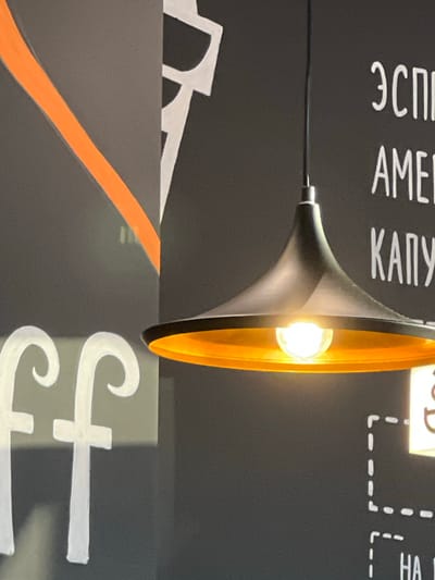 The image shows a close-up of a black wall with white lettering and an orange diagonal stripe. A black pendant light with a bright orange interior hangs in the center, emitting a warm glow from its lit bulb. The lettering on the wall appears to be in Russian, with visible words like "ЭСПР," "АМЕР," and "КАПУ," which likely refer to coffee drinks. On the left side of the wall, two white, stylized letters that resemble "ff" are painted. The background wall is dark gray and slightly textured. There are dashed white lines on the right side of the wall, forming a rectangular diagram, with a partially visible illuminated box above it. The lighting suggests an indoor setting, possibly a cafe or a place related to coffee.