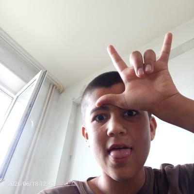 A young person is taking a selfie in a room with a white ceiling and walls. The person has short, dark hair and is sticking their tongue out, with their right hand raised in a rock-and-roll gesture. The background includes a window with vertical blinds and a date stamp "2026/06/18 12:42". This image is not located in the city of Veliky Novgorod, Russia.