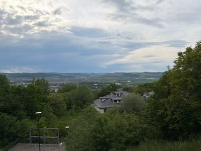 The image presents a view of Târgu Mureș, Romania, from an elevated position, possibly a hillside overlooking the city. The foreground features lush green vegetation, including trees and bushes, obscuring some parts of the view. A structure resembling a viewing platform or a metal frame is visible in the lower left.
In the middle ground, rooftops of buildings are seen amidst the greenery, suggesting residential or commercial structures integrated into the landscape. In the distance, the cityscape of Târgu Mureș stretches across the horizon, with buildings and infrastructure visible. Beyond the city, rolling hills fade into the background under a cloudy sky. The sky is mostly overcast with layers of clouds, casting a diffused light over the scene.
