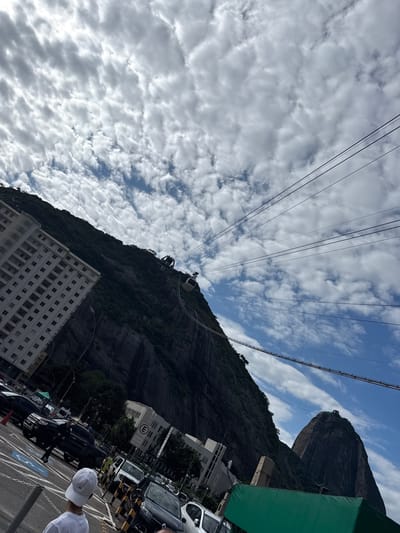 This media file captures a dynamic, tilted upward view of a mountainous landscape integrated with urban infrastructure, characteristic of Rio de Janeiro, Brazil. The scene is dominated by two prominent mountains under a partly cloudy sky during what appears to be daytime.
In the background, a large, dark, forested mountain slopes upwards, its rugged rock face visible. To the upper right, the iconic, distinct peak of Sugarloaf Mountain (Pão de Açúcar) rises against the blue sky. Spanning between these elevated points are multiple strong cables supporting the Sugarloaf Cable Car system. At least two cable cars are visible along these lines, suggesting ongoing operation.
The sky is a mix of scattered, fluffy white and grey clouds (altocumulus or stratocumulus) with patches of clear blue visible through them. The overall lighting suggests bright daylight.
In the foreground and mid-ground, urban elements are visible. A multi-story white building with numerous windows occupies the lower-left portion of the frame. Further to the right, at street level, a parking area with several cars (including white and dark sedans and SUVs) is present. A person wearing a white cap and white shirt is partially visible in the bottom right corner, facing away from the camera. Other pedestrians can be discerned in the background near parked vehicles. A green awning is also partially visible on the far right edge.
Visible text includes a circular sign with a prominent white "E" on a blue background, likely indicating a regulated area such as parking or an entrance. The image's tilted perspective gives a sense of looking up from ground level towards the mountains and the sky.
