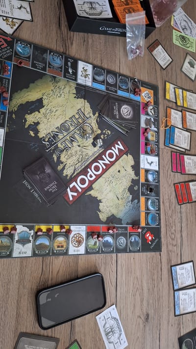 This image shows a Game of Thrones Monopoly board game set up on a wooden surface, likely a table. The game is in progress, with several tokens placed on the board. In the foreground, there are two dice, one showing a 6 and the other a 3, indicating a roll of 9. A stack of "The Iron Throne" cards is also visible on the board.
Surrounding the Monopoly board are various game components. To the left, there is a black box with "GAME OF THRONES" printed on it, along with stacks of money cards, some of which are orange and marked with "500". There are also some pink and white cards scattered around. A small plastic bag containing red tokens is also present.
To the right of the Monopoly board, a smartphone with a black case is placed on the table. A Monopoly money bill, featuring the Game of Thrones logo, is also visible. Further to the right and slightly out of focus, there is a green card and another card with text on it.
The game pieces and board suggest a focus on the Game of Thrones universe, with locations and houses from the series represented. The arrangement of the game components indicates an active gameplay session. The lighting suggests it's daytime, and there are no indicators of weather or specific emotions. The wooden texture of the surface provides a rustic feel. The visible text includes "MONOPOLY", "GAME OF THRONES", "THE IRON THRONE", and denominations on the money cards.