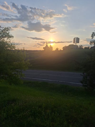 The image depicts a rural landscape at sunset. A road with two lanes and white markings cuts horizontally across the mid-section of the frame. On the left side of the road, lush green grass with small pink flowers covers the foreground. Further up, a tree with vibrant green leaves stands prominently. On the right side of the road, the terrain slopes upwards, covered in grass. In the distance, silhouetted against the setting sun, is a small church with a cross on its roof. The sun is a bright orb, casting a warm glow and creating sunrays that extend across the sky. The sky itself is a mix of pale blue and scattered clouds, some of which are illuminated by the sunset. A modern, square billboard structure is visible on the right side of the hill, near the horizon, along with a street lamp. The overall scene suggests a serene and picturesque evening in a rural setting, potentially near the city of Kapyĺ, Belarus, given the architectural style of the church and the general landscape.