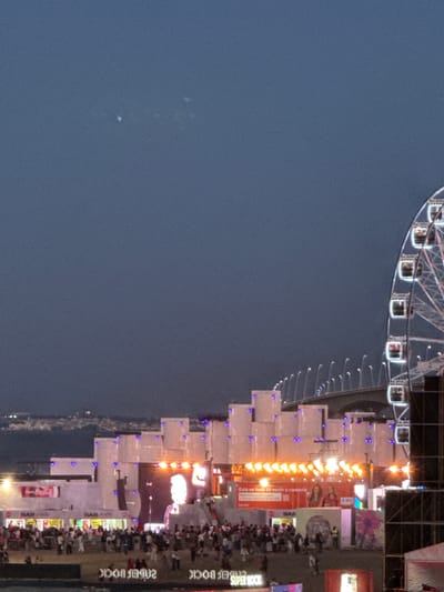 The image depicts the Super Bock festival, a music event held in Parque das Nações, Lisbon, Portugal. In the foreground, a crowd of people is visible, with some individuals in the middle ground standing near a stage with bright lights and a large screen displaying an image. To the right, a Ferris wheel with lit cabins is partially visible, and a bridge spans across the background. The sky is dark, indicating it is likely evening or night. The overall scene captures the lively atmosphere of a large outdoor festival.