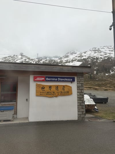 The image shows an outdoor scene near the Bernina Diavolezza railway station, located in Pontresina, Switzerland, with a snowy mountain visible in the background under a cloudy sky. The station building has a light-colored facade with a sign that reads "Bernina Diavolezza" and another wooden sign written in Japanese, referring to the Albula/Bernina Lines as a World Heritage Site. There is a stone wall next to the building and a paved road in the foreground. A bin is located on the side.