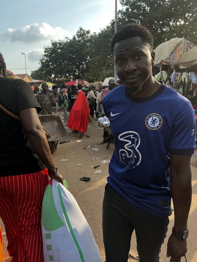 In Nyanya, Nigeria, a man wearing a blue Chelsea Football Club jersey smiles at the camera. He is standing in what appears to be a busy outdoor market. In the background, numerous people are visible, along with market stalls and various goods. A large red cloth is draped over something in the middle ground. The ground is a dirt or paved surface littered with debris. The sky is partly cloudy and the lighting suggests it is daytime. The man is wearing a watch on his left wrist. In the foreground, another person's arm is visible, holding a white and green patterned pillow. The overall scene depicts a lively marketplace environment.