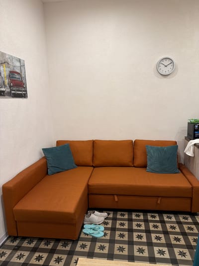 An interior scene depicts a corner of a room with a rust-colored sectional sofa positioned against a textured, off-white wall. Two teal throw pillows rest on the sofa. Above the sofa, a framed picture of a vintage red car is mounted on the wall. To the right of the sofa, a microwave oven is visible on a shelf, with a folded white towel hanging to its left. On the floor in front of the sofa, a pair of light blue flip-flops and a pair of grey slip-on shoes are placed. The floor is tiled with a repeating black, grey, and beige geometric pattern. A round clock with black numbers and hands is mounted on the wall above the sofa. The overall impression is that of a living space or a small apartment. This scene is in the city of Catania, Italy.