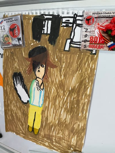 This image depicts a drawing on a whiteboard, likely in Maksimkovo, Russia, given the presence of Russian text on some of the attached items. The central subject is a child's drawing of a person with brown hair, a black beret, a striped shirt, and yellow pants. The figure has a distinctive dark shadow over its face. To the left of the drawing is a small square item with the text "ИДИ ВПЕРЕД И НЕ СДАВАТЬСЯ" which translates to "Go Forward and Don't Give Up". It features a karate emblem and a picture of a person in a space helmet. To the right is a rectangular item with Russian text that reads "ВЕЧНАЯ СЛАВА ГЕРОЯМ ВЕЛИКОЙ ПОБЕДЫ" which translates to "Eternal Glory to the Heroes of the Great Victory". This item also shows a depiction of Victory Day celebrations with red carnations, a statue of a woman, and the numbers "20 25 80 ПОБЕДА!" (2025 80 Victory!). The drawing is rendered in bold, sketchy strokes with brown and gold markers, and the figure appears to be standing in a textured environment. The overall scene is informal, suggesting a home or school setting where children's artwork and commemorative items are displayed.