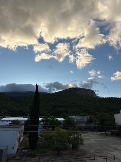 The image captures a natural landscape with some man-made structures in the foreground. Dominating the upper portion of the frame are dramatic, scattered clouds against a bright blue sky. The clouds are a mix of bright white and darker grey, suggesting varying levels of thickness and light penetration. Below the sky, a lush, dark green forested mountainside slopes upwards, culminating in a rocky, lighter-colored peak on the right. A solitary, tall, dark green cypress tree stands prominently in the left middle ground, its slender form contrasting with the broader shapes of the mountain. In the lower portion of the image, several utilitarian, low-rise buildings are visible, characterized by white facades and flat, light-colored roofs. A large, paved, somewhat cracked area, possibly a courtyard or parking lot, occupies the foreground. The overall scene suggests an outdoor setting with a blend of natural and industrial or institutional elements, possibly in a semi-urban or exurban area. The location in Chaika, Ukraine is not visually indicated by any specific landmarks or signage within the image. There are no people or discernible actions depicted.