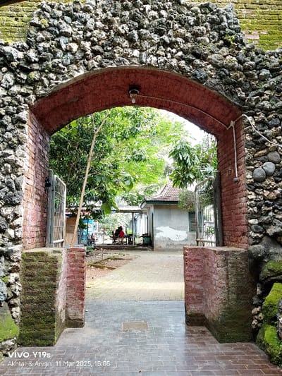 The image shows a brick archway with a red arch, made from irregularly shaped stones, leading to a courtyard at the location of Keraton Kasepuhan, Cirebon, Indonesia. There is a building visible behind the archway. The courtyard is paved with gray brick. Two people can be seen sitting at a table in the background. The archway is surrounded by vegetation, including moss and trees. The building is gray, with a wooden door and windows. There is a fence and another gate behind the archway. The light source is an overhead light inside the archway. The image is taken from a low angle, looking up towards the archway.