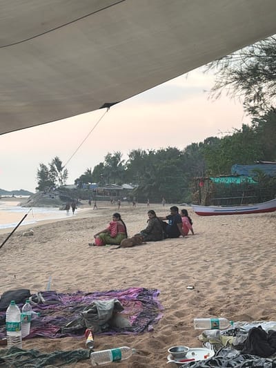 The image depicts a sandy beach scene during what appears to be dusk or dawn, characterized by a light pink and orange sky. The immediate foreground is partially obscured by a light-colored canvas or tarp stretched overhead, from which a rope descends towards the lower left.

On the sand directly below the tarp, a multicolored fabric (purple and black patterns) is laid out. On or near this fabric are two Bisleri brand plastic water bottles, one mostly full and one partially filled, a crushed red and white aluminum can, and a small, light brown puppy resting. To the right, another empty plastic bottle, a white plate with food remnants and a small metal bowl, and various discarded dark fabrics are visible on the sand. A light-colored, rectangular object rests on the sand further to the right.

In the midground, four individuals are seated on the sand, facing towards the left. A woman in a green and pink saree is on the left, with a light brown dog resting beside her. Next to her are a woman in a dark patterned outfit, a man in a dark t-shirt, and another woman in a pink top. Further left, closer to the water, a white dog is lying on the sand. A dark pole extends upwards from the lower left, partially visible.

The background reveals a broader view of the beach. On the left, the ocean's edge shows gentle waves breaking against a rocky outcrop, upon which several individuals are standing. Numerous people, including adults and children, are scattered along the beach and at the water's edge, engaged in various activities like walking and playing. Towards the right, a white and red fishing boat is beached on the sand, adjacent to dense green foliage, behind which several rustic structures, some covered with blue tarps, are nestled. This scene is identified as taking place in Gokarna, India.