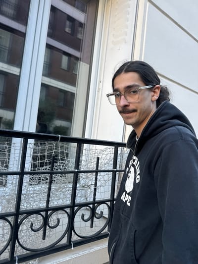A male subject is depicted in a medium shot, positioned on what appears to be a balcony or ledge in Vincennes, France. The subject has dark, medium-length hair styled back, a thin mustache, and wears clear-framed eyeglasses. He is wearing a dark, zip-up hooded sweatshirt featuring a white graphic logo of a gorilla head with the text "BATHING APE". The subject's head is slightly turned, and he is looking directly at the camera with a neutral expression.
In the foreground, a black wrought-iron balcony railing with decorative elements is present. Attached to this railing is a white, wide-mesh netting, secured with white zip ties, spanning across the visible section of the railing.
The immediate background shows a white building facade with a tall, white-framed window. The window's surface reflects an opposing multi-story building, characterized by multiple rectangular windows and a dark, possibly reddish-brown, facade. A faint reflection of greenery, possibly a tree, is also visible within the window pane.