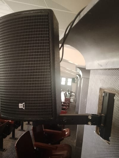 The image shows a close-up of a black speaker mounted on a metal bracket, attached to a perforated wall. The speaker has a grid-like texture on its front and a logo that appears to be "CK" with an arrow. In the background, rows of empty seats with red upholstery are visible, suggesting an auditorium, theater, or classroom setting. Sunlight streams in from windows at the far end of the room. The overall environment appears to be indoors, with a textured ceiling. The image does not provide any information to determine its location in Kuson, Nigeria.