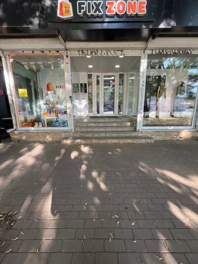 The image shows the storefront of "FIX ZONE", a store located in Voronezh, Russia. The storefront features a large, illuminated sign that reads "FIX ZONE" in orange lettering. The store has large glass windows displaying merchandise. To the left, the window showcases stuffed animals and various products. To the right, the window displays clothing and mannequins. The entrance to the store is in the center, with a set of stairs leading up to it. The building is situated on a brick-paved sidewalk, with shadows from trees cast across it. The overall scene is that of a commercial street in a city.