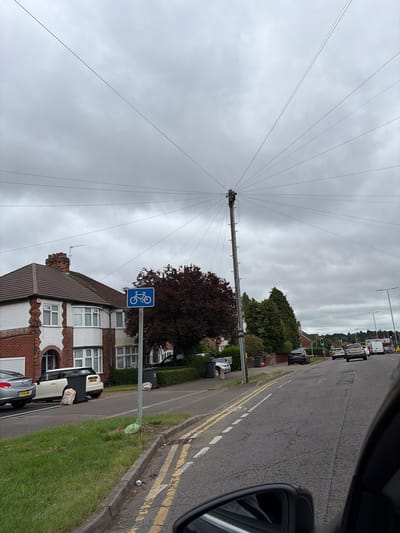 The image captures an overcast daytime scene on a residential street, likely in Leicester, United Kingdom. The photograph is taken from inside a vehicle, looking towards the passenger side and front.
A tall, weathered utility pole dominates the midground, positioned on the left side of the road. Numerous dark wires extend outwards from its top, fanning across the uniformly grey, cloudy sky, suggesting it serves as a central hub for telecommunications or power distribution. A person, possibly a utility worker, is discernible near the very top of the pole, engaged in work on the lines.
To the right, a tarmac road stretches into the distance, with a clear yellow double line marking the curb, typical of UK road regulations prohibiting parking. Several vehicles are visible; some are parked along the pavement, while others are seen driving further down the road. On the pavement to the left, a blue square sign with a white bicycle symbol indicates a designated cycle route or facility.
Houses with traditional British architecture, featuring brickwork, bay windows, and tiled roofs, line the street behind the pole and various trees, including one with dark foliage. A neatly maintained grassy verge runs alongside the road, separating it from the pavement. The overall scene depicts a functional urban environment under a flat, grey sky, with everyday activity unfolding. No specific emotions are conveyed.
