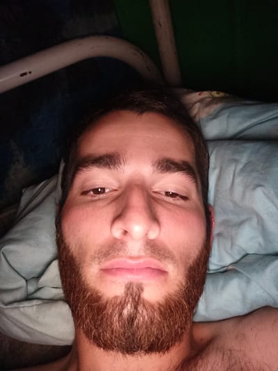 The image is a close-up, overhead shot of a man lying down, possibly in bed, with his face filling most of the frame. He has brown hair and a neatly trimmed beard. His eyes are open, and he is looking directly at the camera. The background consists of a light blue pillow and a dark green surface. There is also a glimpse of a metal bar in the upper part of the image. The man appears to be shirtless. The scene is indoors, with low lighting conditions.