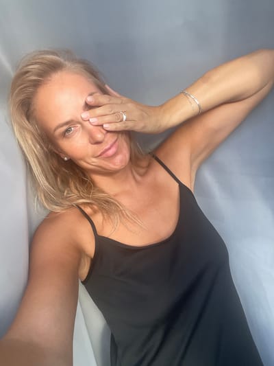 A blonde woman with her right hand covering her right eye smiles at the camera, with her left arm extended outwards. She is wearing a black tank top and a silver bracelet on her left wrist. The background is a greyish-blue fabric. The image is taken from the shoulders up. There is no information to suggest this image is located in Baranovka, Russia.
