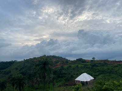 The image shows a hilly landscape in Asaga Amangwu, Nigeria. Overcast skies filled with thick, gray clouds dominate the top half of the frame. A partially constructed building with a white roof is visible in the lower right foreground. The hillside is covered in lush green vegetation, including palm trees. Evidence of erosion is visible as exposed soil on the hillside.