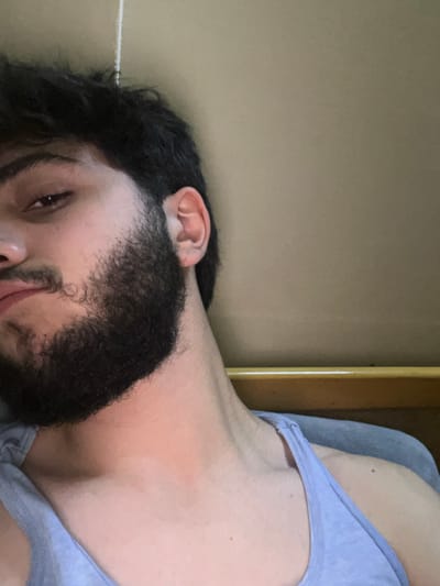 A man with a dark beard and wavy dark hair is lying down, his face tilted toward the camera. His ear is visible against a beige wall. He is wearing a light blue tank top, and part of his neck and shoulder are visible. The background suggests he is indoors, possibly in a room with light-colored walls.