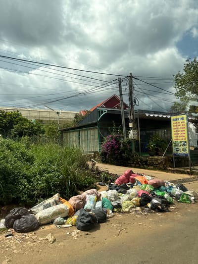 A pile of trash bags and loose garbage is accumulated on the side of a road in Thôn 1, Vietnam. The garbage includes black plastic bags, sacks of what appears to be cement or fertilizer, and various colorful plastic bags containing refuse. Behind the trash, there is a building with a green metal facade and a red roof, suggesting it might be a commercial establishment, possibly a restaurant, as a yellow sign with Vietnamese text is visible. Lush green vegetation and trees surround the building. Overhead, a complex network of electrical wires crisscrosses the sky, which is filled with heavy, gray clouds. In the distance, a greenhouse-like structure is visible, and a small wind turbine stands next to it. The road appears to be paved with asphalt and shows signs of wear. The scene suggests a rural or semi-urban area.