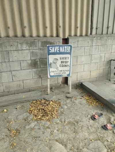 The image shows a sign attached to a metal pole in front of a brick wall. The sign reads "Save Water, Every Drop Counts, Turn Taps Off Properly & Report Leaks". It is located in Ashaka, Nigeria at latitude: 10.929, longitude: 11.475. On the ground in front of the sign is a pile of dried leaves. There are two pairs of sandals on the ground to the right of the sign. The environment is dry and dusty.
