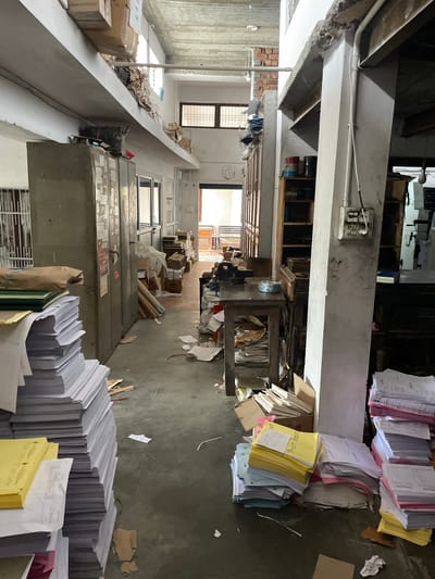 The image depicts a cluttered interior space in what appears to be an office or storage area. Piles of paper, files, and boxes are scattered throughout the scene. Furniture, including metal cabinets and shelves, is visible amidst the clutter. The scene is situated within a city, specifically Dharamshala, India. Natural light enters through windows at the far end of the passage, but the overall atmosphere is somewhat dimly lit and disordered. There are no people visible in the frame.